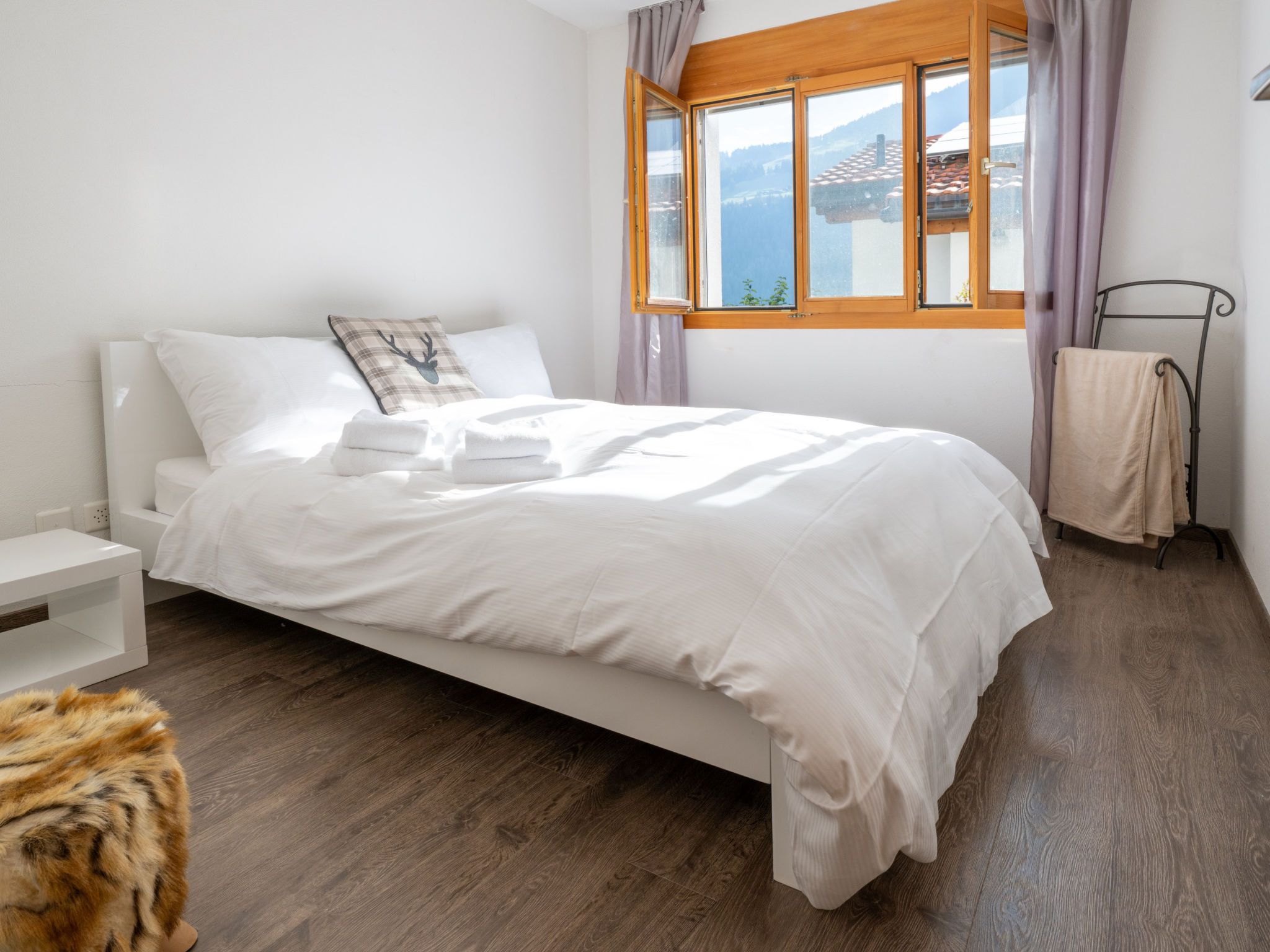Ein ordentlich bezogenes Bett mit weißen Leinen und einem Kopfkissen mit Hirschmuster, ein Nachttisch, ein Fenster mit Bergblick und ein Fellteppich auf dem Boden.