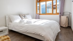 Een netjes opgemaakt bed met witte lakens en een sierkussen met een hertenmotief, een nachtkastje, een raam met uitzicht op een berg en een bontje op de vloer.