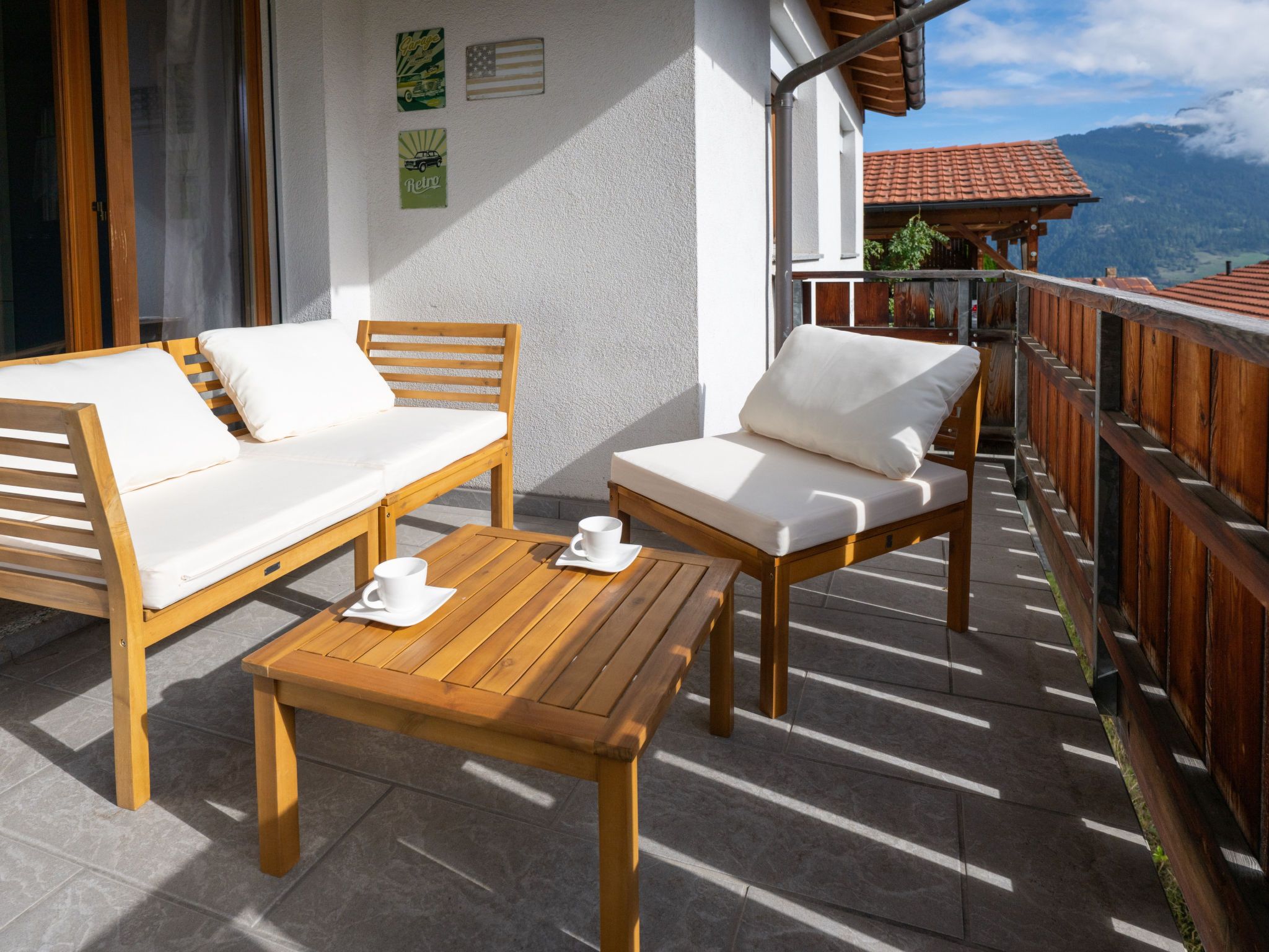 Un balcón con una mesa de madera y dos sillas blancas con cojines, una valla de madera y vistas a la montaña.