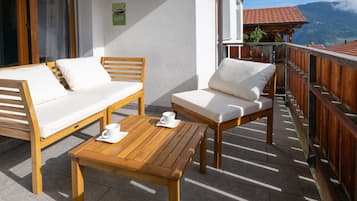Un balcón con una mesa de madera y dos sillas blancas con cojines, una valla de madera y vistas a la montaña.