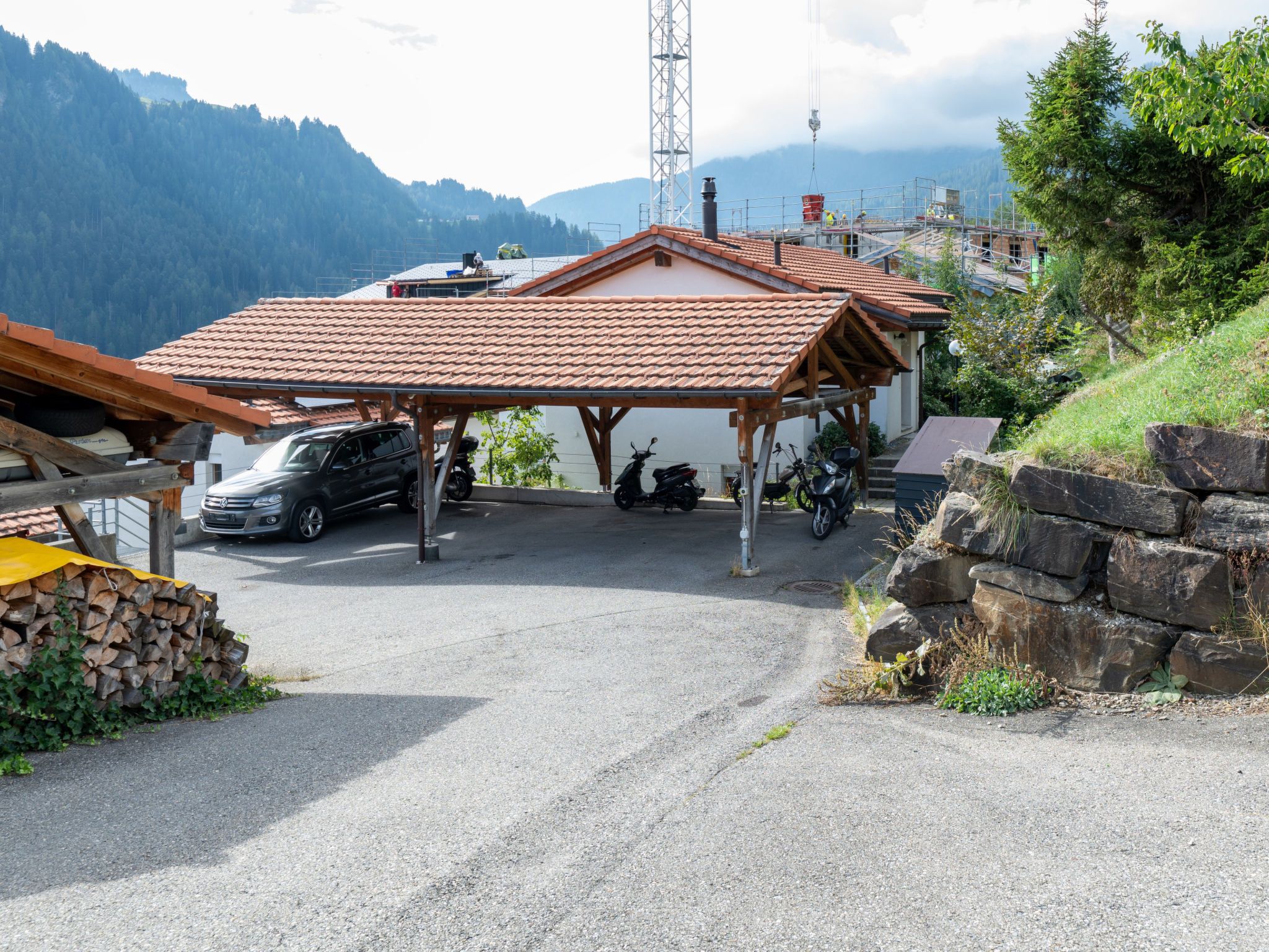 Un área de estacionamiento techada con un coche y una moto, rodeada por un paisaje montañoso.
