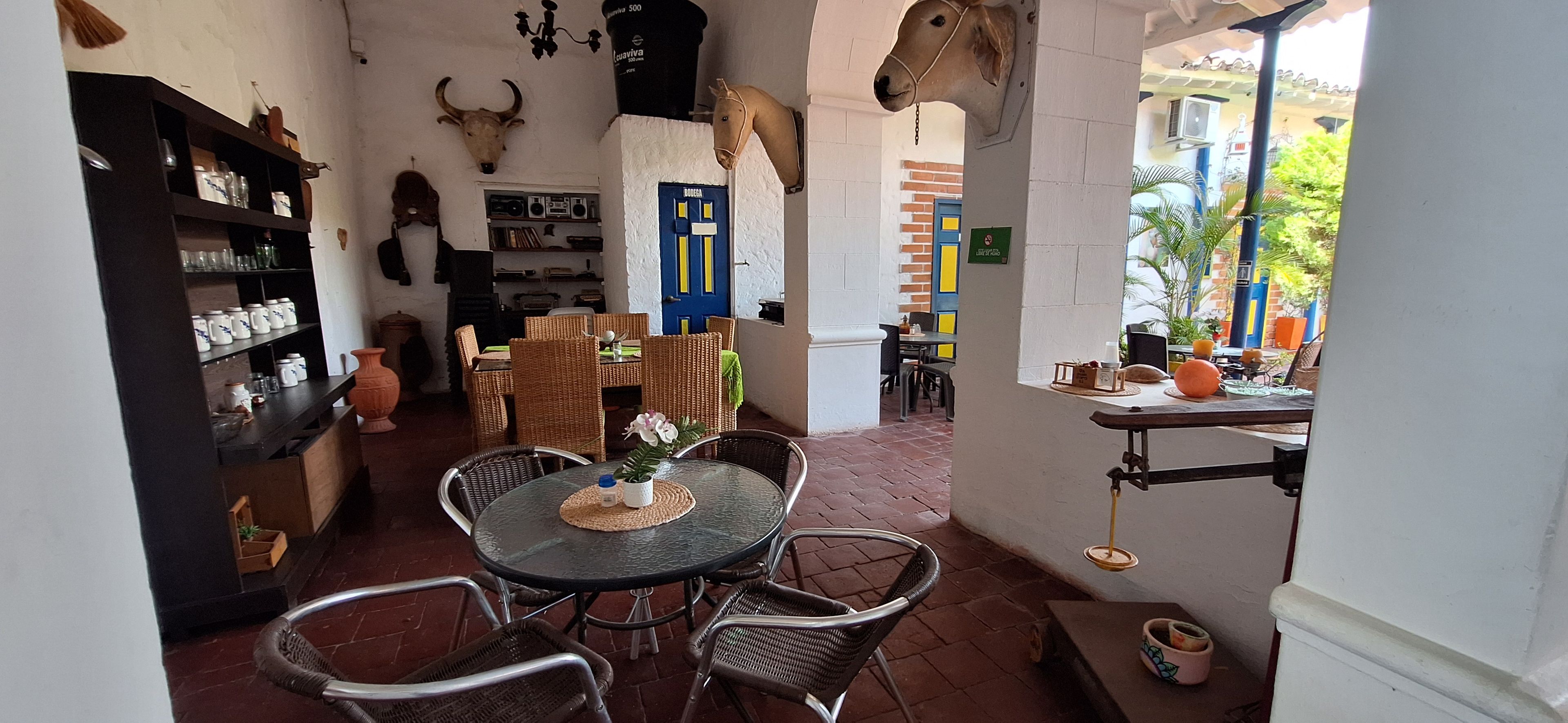 Un interior acogedor con una zona de comedor que incluye sillas de mimbre y una mesa de vidrio, una cocina con estantes y utensilios, y una puerta azul.