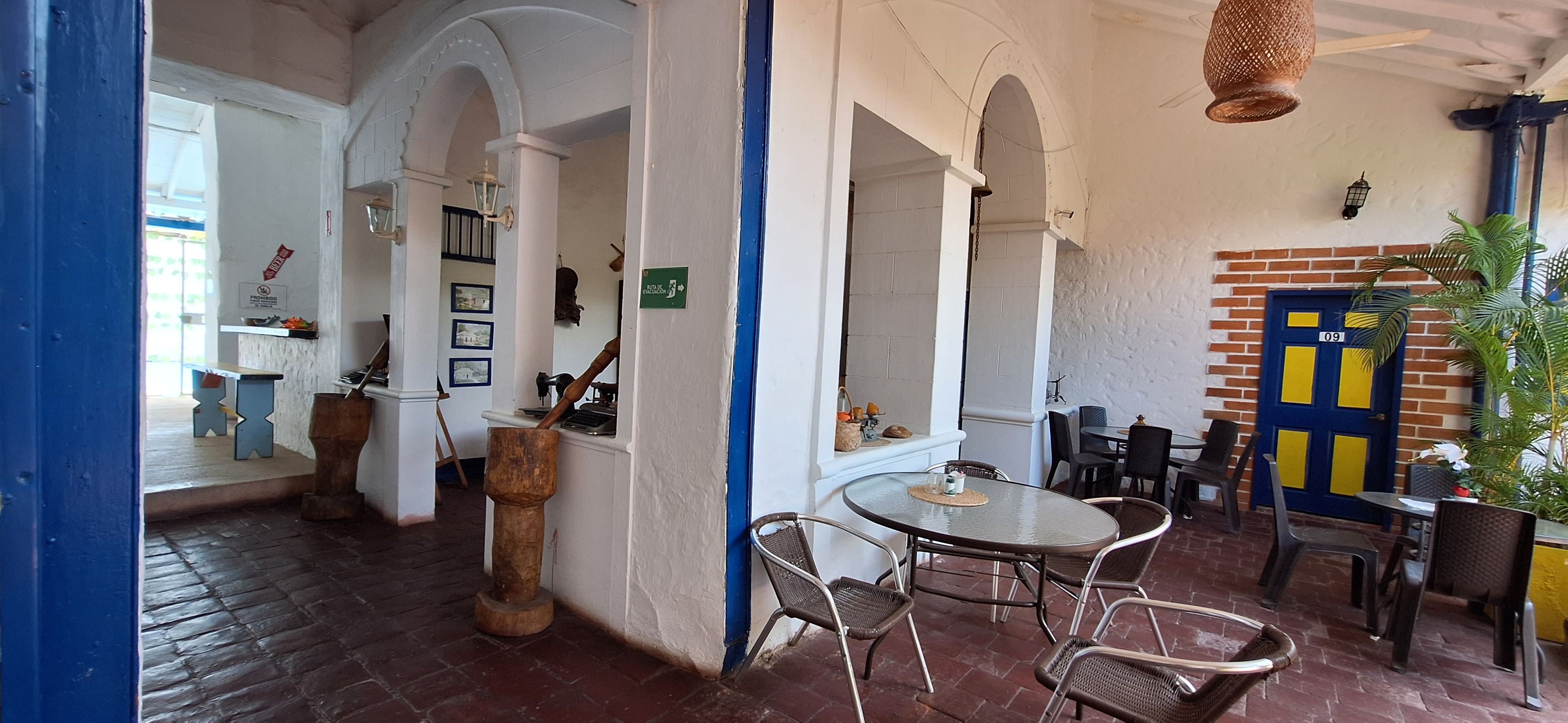 Restaurante com chão de tijolos, paredes brancas e detalhes em azul. Há mesas e cadeiras para refeições.