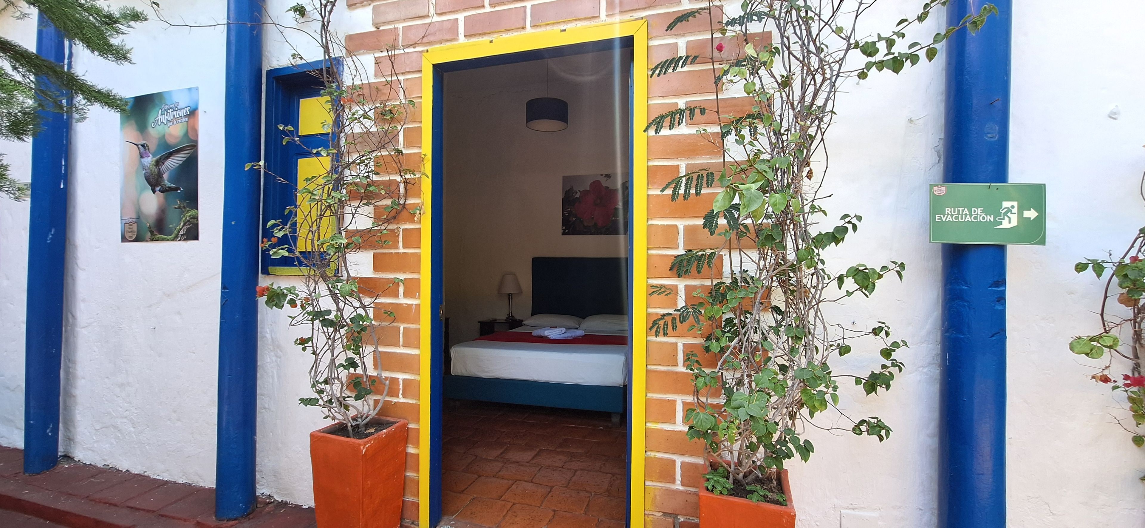 Uma porta dá acesso a um quarto com uma cama, mesa de cabeceira e luminária na parede. Há uma placa verde indicando uma rota de fuga.