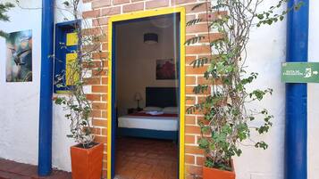 Uma porta dá acesso a um quarto com uma cama, mesa de cabeceira e luminária na parede. Há uma placa verde indicando uma rota de fuga.