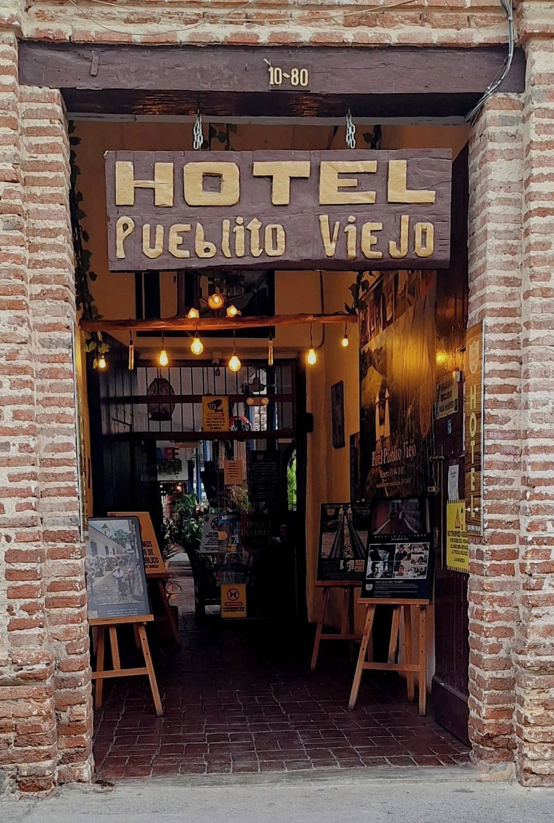 De ingang van een hotel met een rustieke baksteenvoorgevel, een bord waarop 'HOTEL Pueblo Viejo' staat en een houten schildersezel met een kaart of poster ernaast.