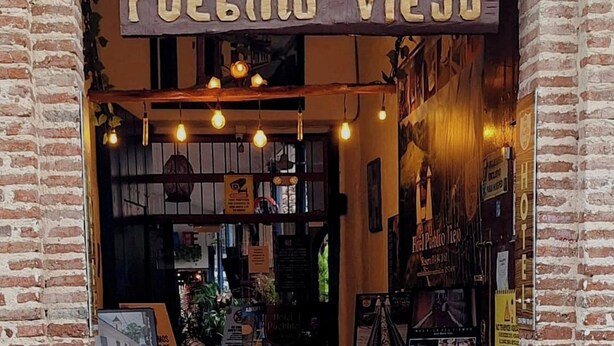 L'ingresso di un hotel con facciata in mattoni rustici, un cartello che riporta 'HOTEL Pueblo Viejo' e un cavalletto di legno con una mappa o un manifesto di fronte.