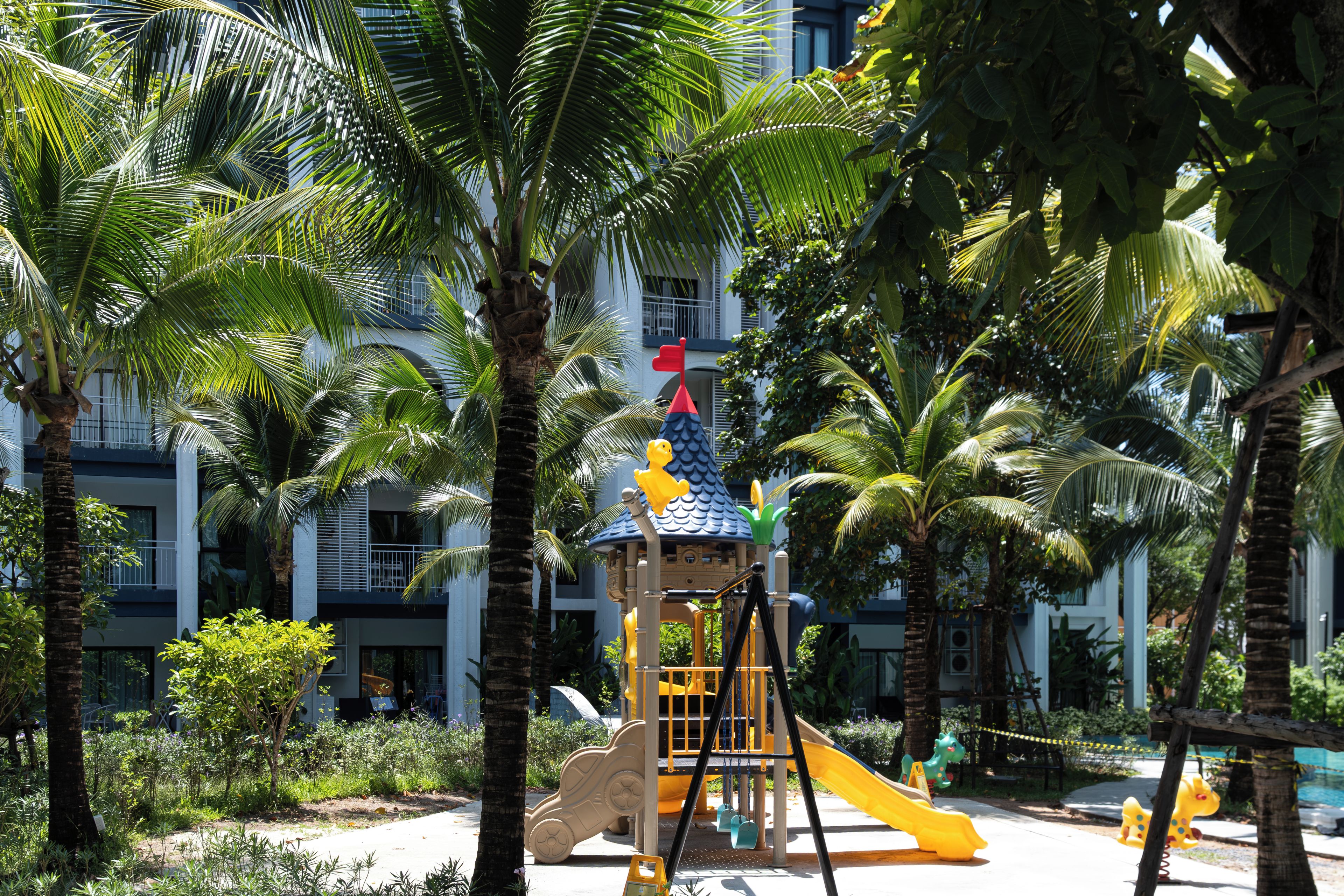 A playground with a slide and climbing structures, surrounded by palm trees and a building in the background.