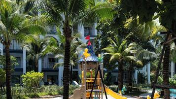 A playground with a slide and climbing structures, surrounded by palm trees and a building in the background.