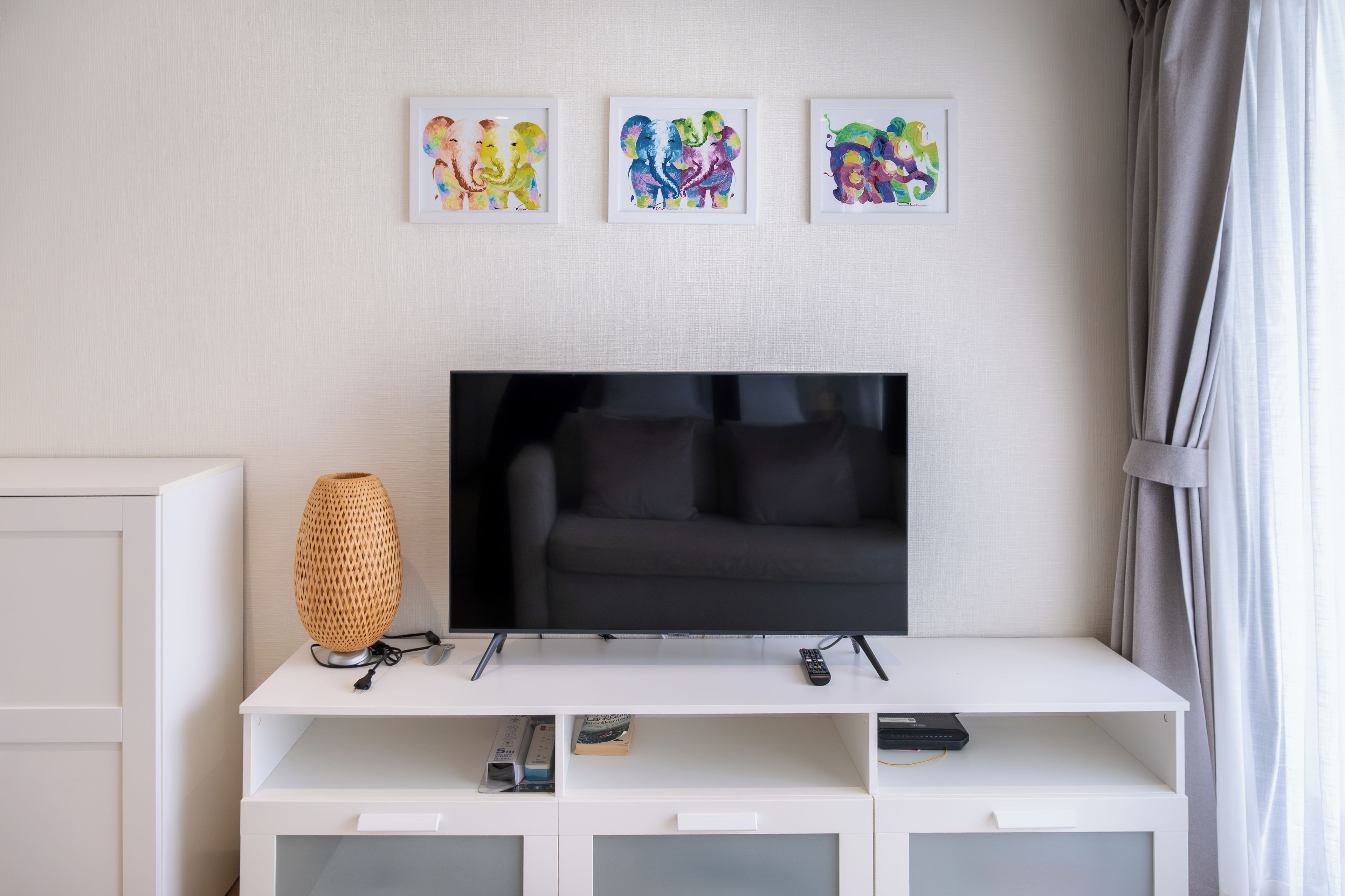 A modern living room with a flat-screen TV on a white entertainment unit, three framed artworks above the TV, and a woven lamp on the left.