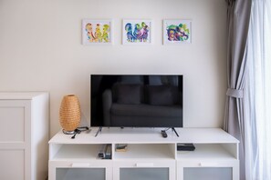 Uma sala de estar moderna com uma TV de tela plana sobre um móvel branco, três obras de arte emolduradas acima da TV, e uma luminária trançada à esquerda.