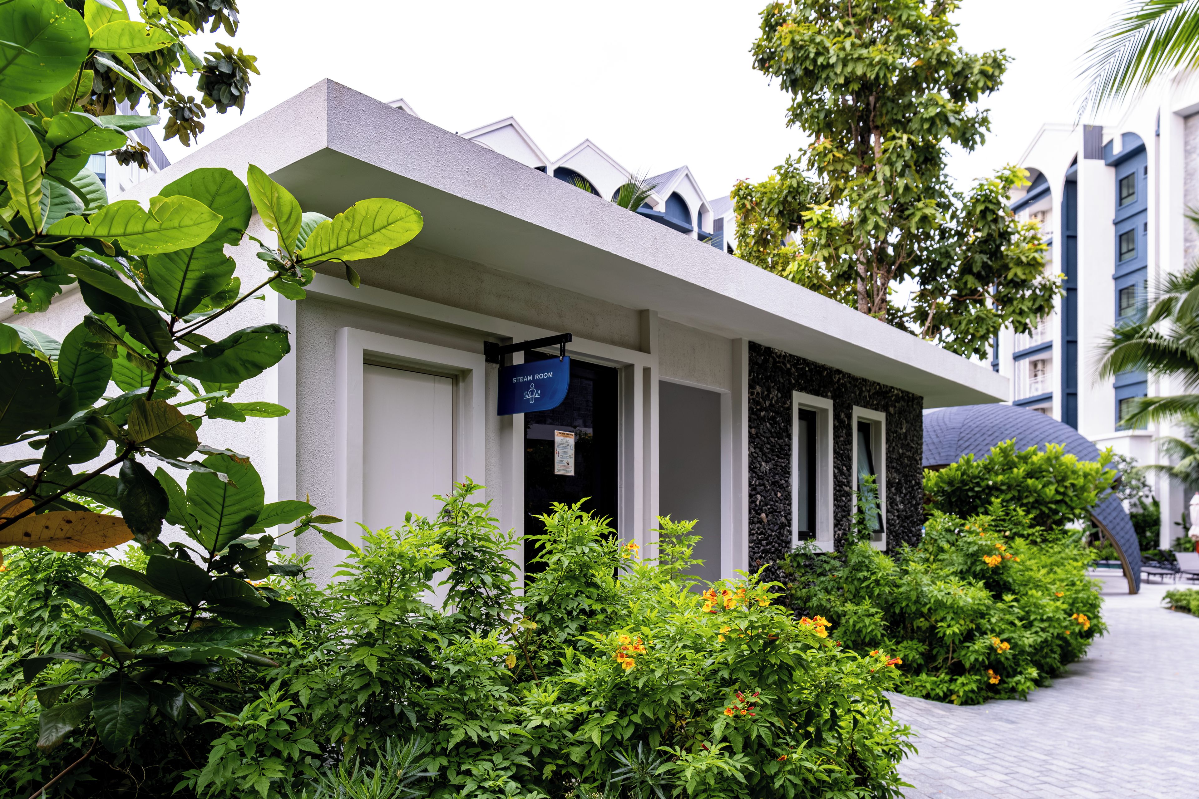 A modern hotel with a clean, white facade, a sign indicating a steam room, and lush greenery surrounding the building.