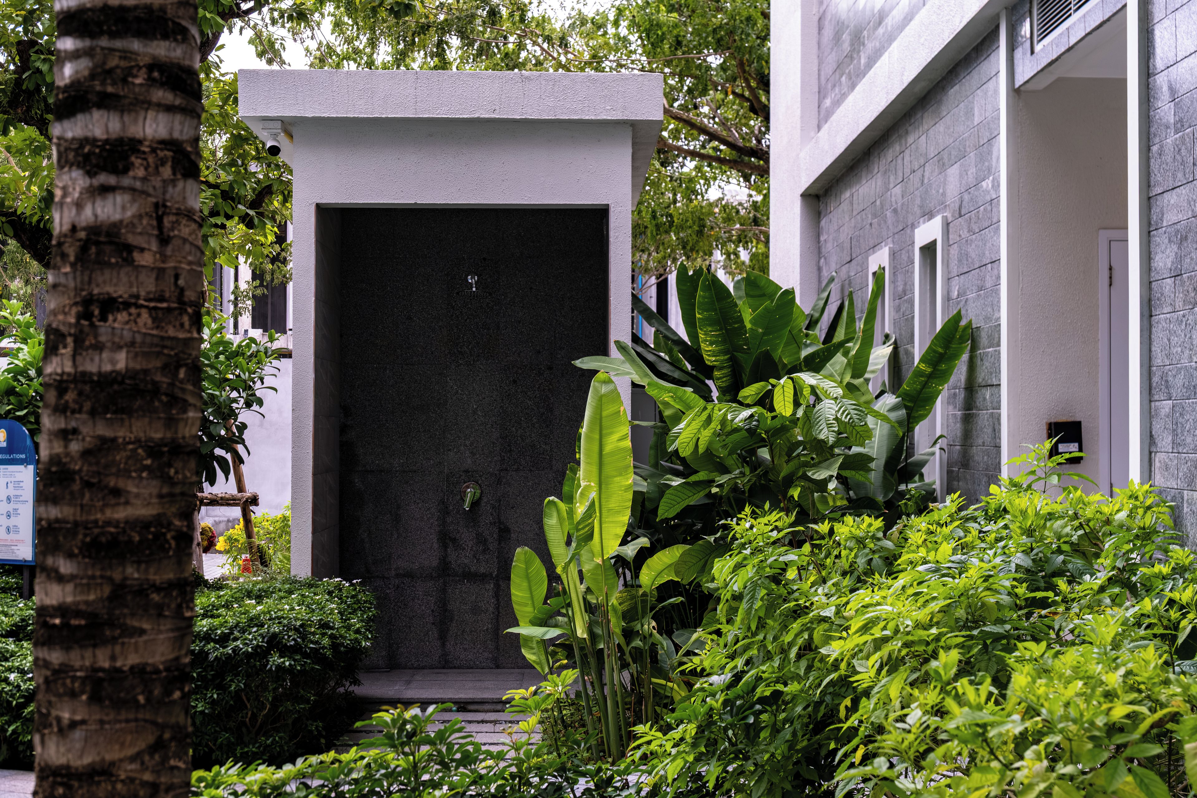 A building entrance with a dark facade, surrounded by lush greenery and a palm tree.