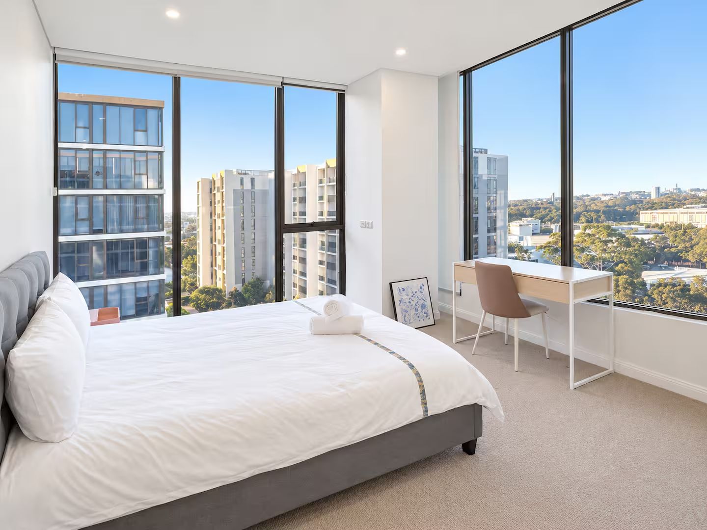 A modern bedroom with a large bed, a desk, and a view of the cityscape.