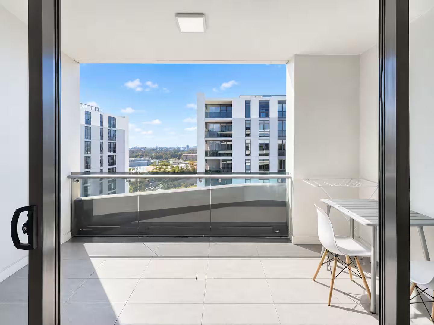A modern balcony with a clear view of surrounding buildings, a glass door, and a small dining area with chairs.