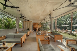 A covered outdoor seating area with multiple tables and chairs, a wooden wall, and a view of a pool and greenery. - Saneca Resort Ubud (Ubud)