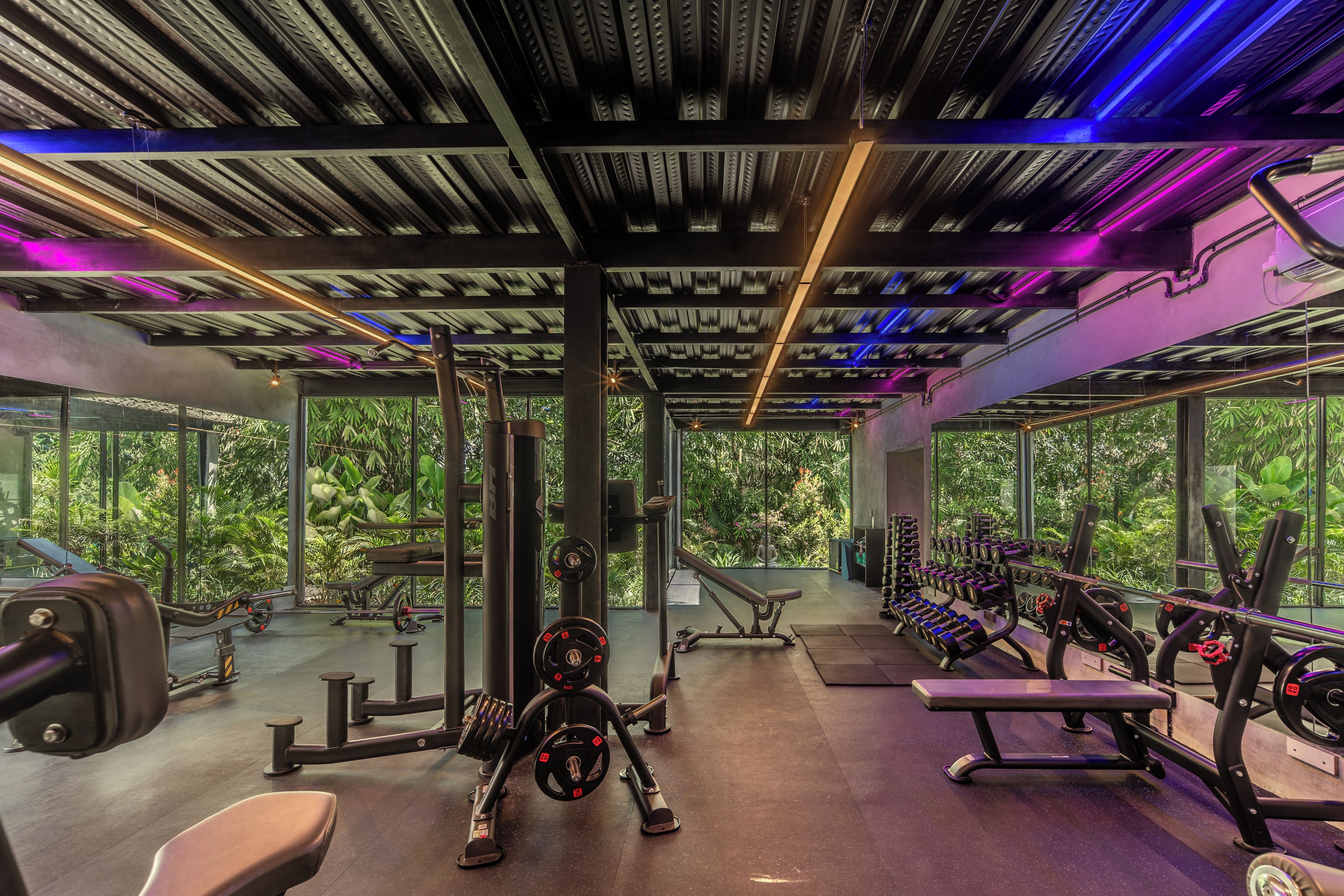 Un gimnasio con diverso equipo de ejercicio, ventanas amplias y un techo con vigas y luces a la vista.