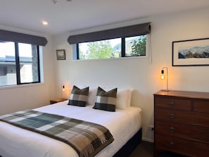 Ein Schlafzimmer mit Bett, Kommode, Nachttisch, Lampe und einem Fenster mit Blick.
