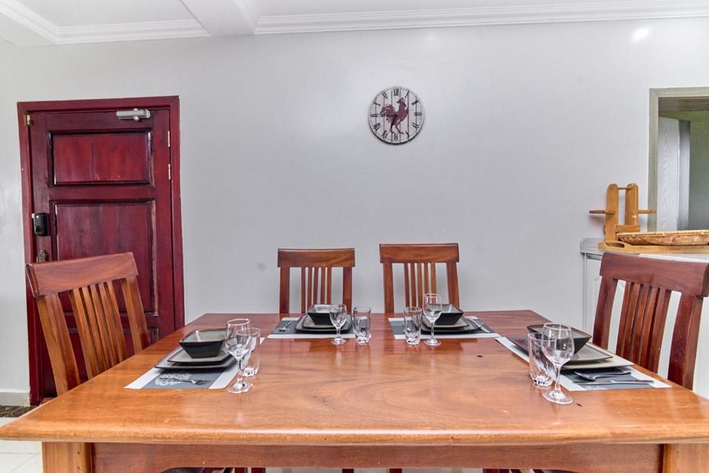 Un comedor con una mesa de madera dispuesta para cuatro personas, con dos sillas a cada lado y un reloj fijado a la pared.
