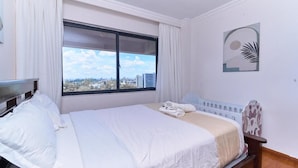 A hotel room with a bed, a window with curtains, a view of buildings, and a wall with a decorative piece. - Crestpark Skyline Penthouse (Nairobi)
