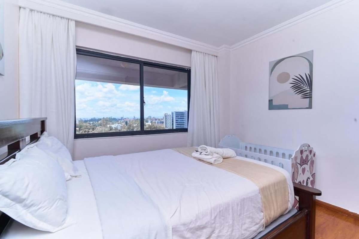A hotel room with a bed, a window with curtains, a view of buildings, and a wall with a decorative piece.