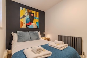 A hotel room with a bed, a blue blanket, two towels, a painting on the wall, a bedside table with a lamp, and a radiator. - Bright 2BR Oxford Flat | Free Parking & City Access (Oxfordshire)