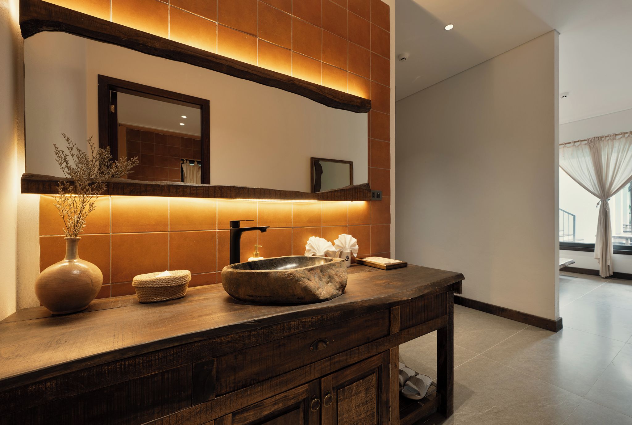 A modern bathroom with a wooden vanity, a large mirror, and a vase with dried flowers.