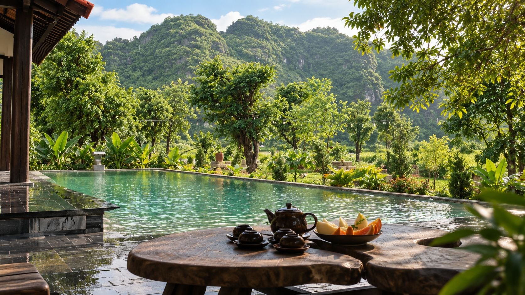 A serene pool area with a table set for tea and fruit, surrounded by lush greenery and mountains.