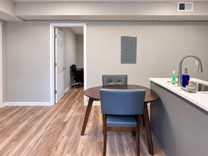Un coin repas avec une table ronde et deux chaises, une cuisine équipée d’un évier et d’un robinet, et une porte donnant sur une autre pièce.