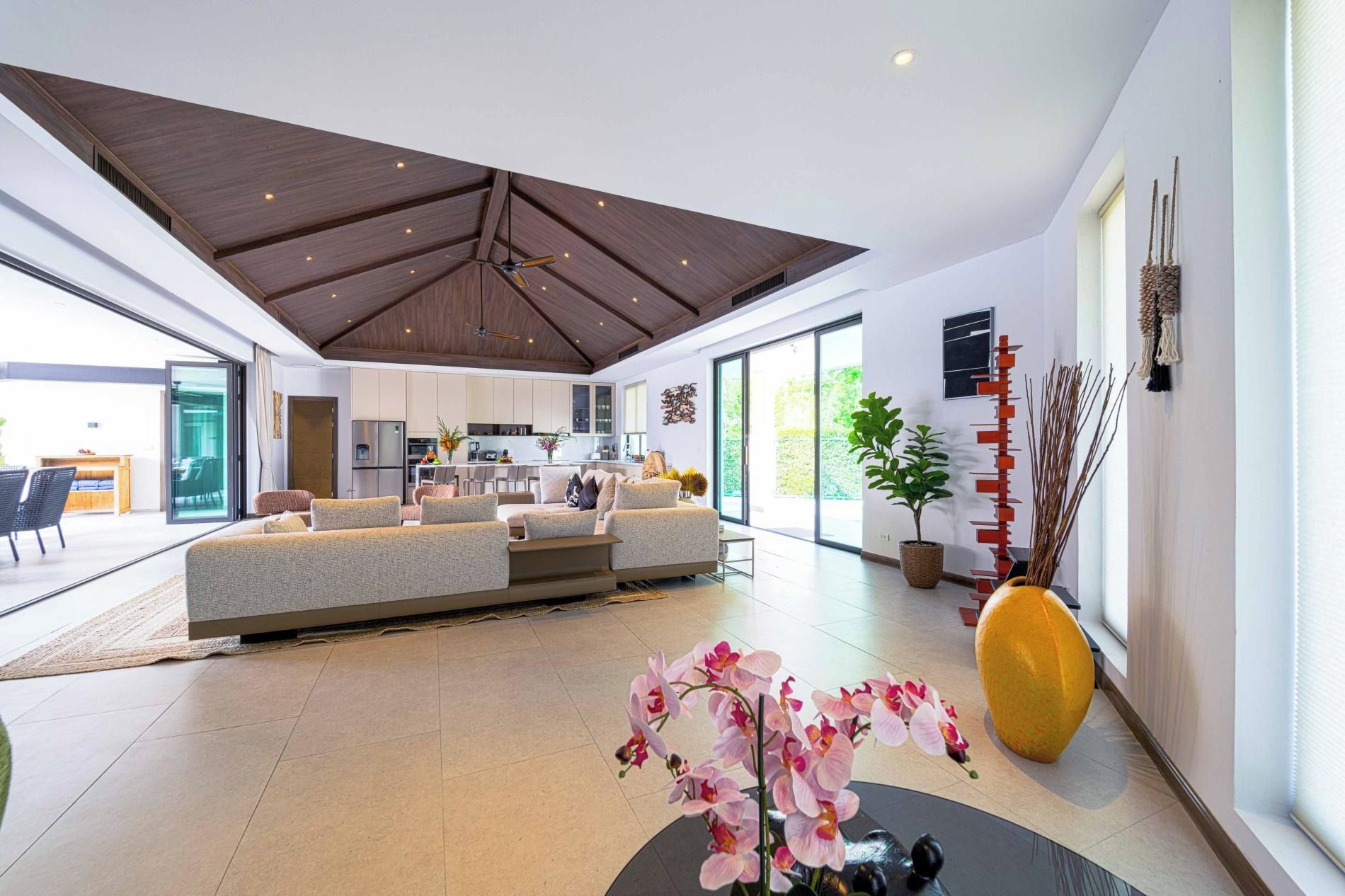 A spacious living area with a high ceiling, a sofa set, a dining area, and a large window providing natural light.