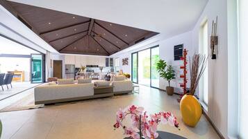 A spacious living area with a high ceiling, a sofa set, a dining area, and a large window providing natural light.