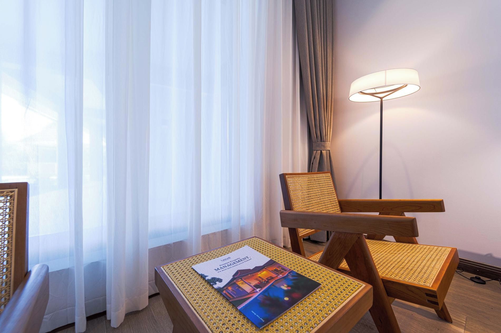 Une chambre d’hôtel avec un lit, une chaise, une table avec un magazine, une lampe sur pied et des rideaux légers.