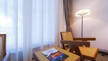 Une chambre d’hôtel avec un lit, une chaise, une table avec un magazine, une lampe sur pied et des rideaux légers.