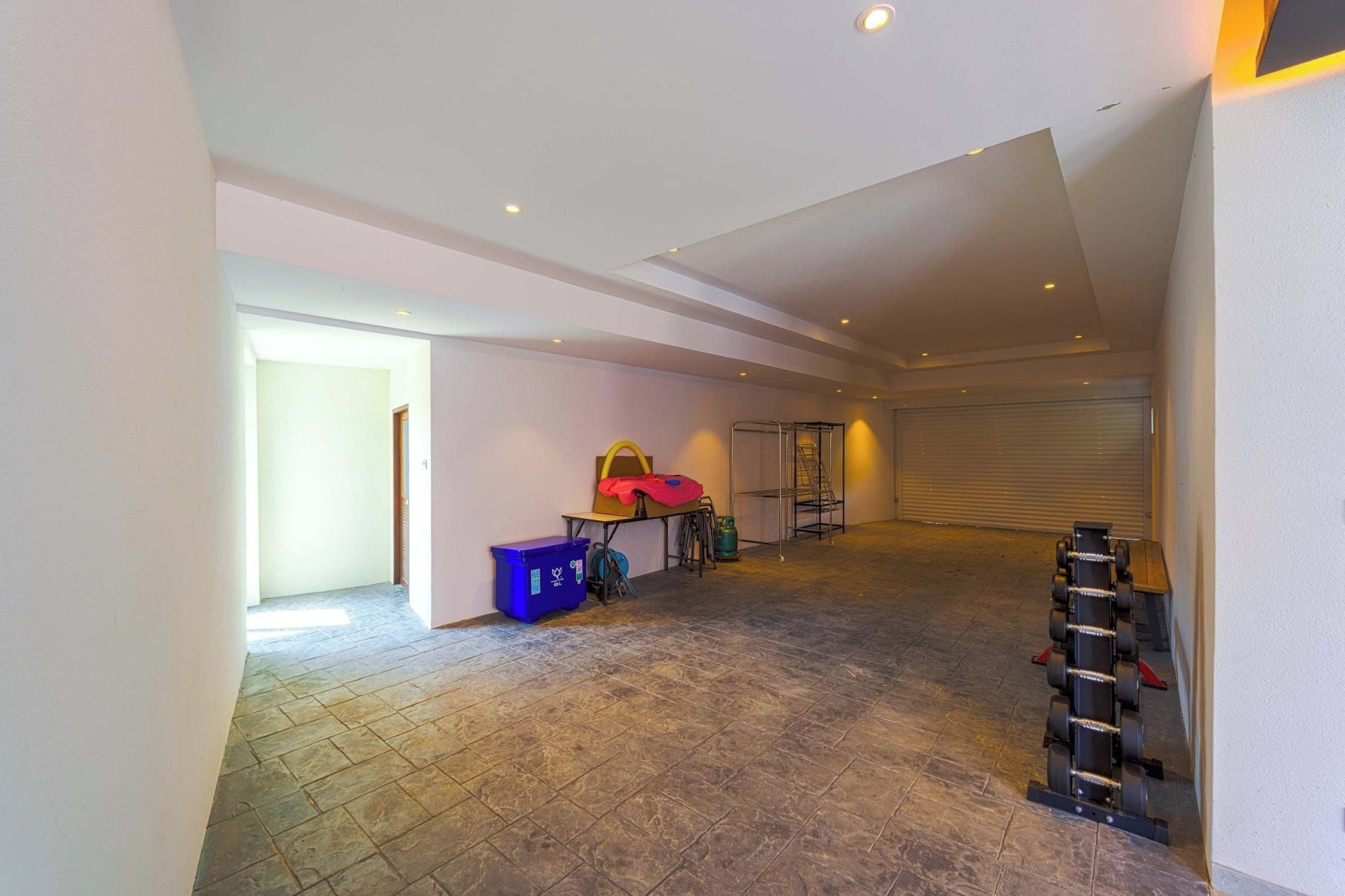 A spacious, well-lit room with a tiled floor, a metal rack with weights, and a blue trash bin.