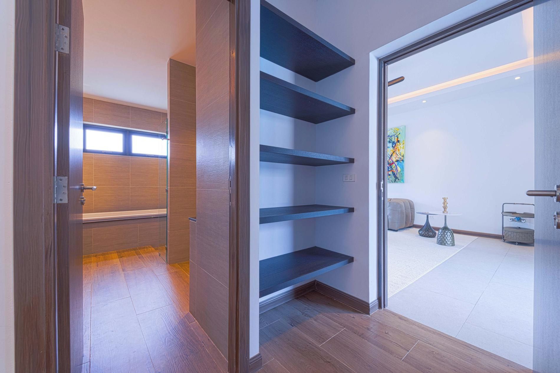 Un pasillo con suelo de madera, una puerta de vidrio que da a una habitación con una estantería azul y un cuadro en la pared.