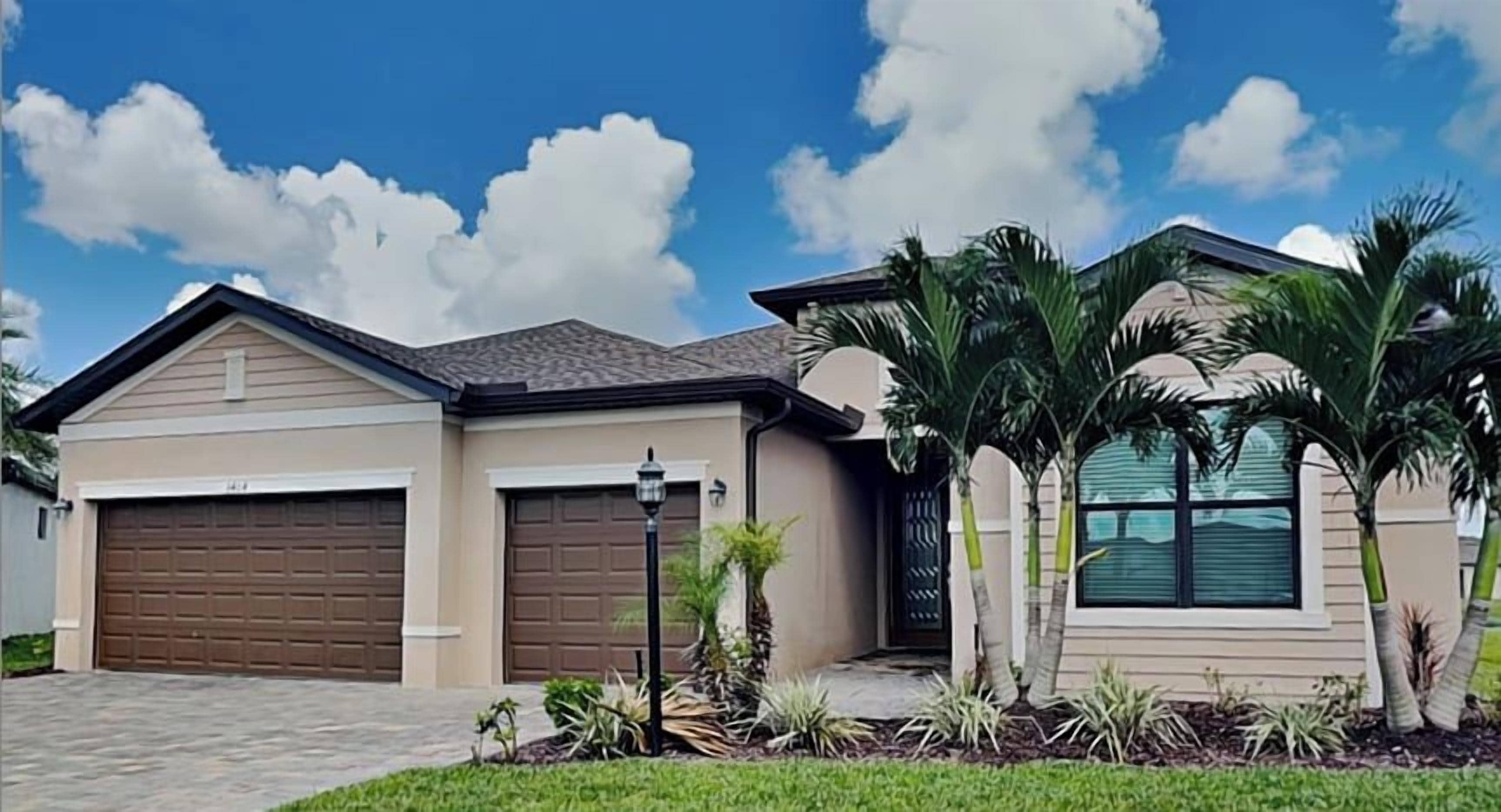 A residential house with two garage doors, a paved driveway, and a well-maintained lawn with palm trees.