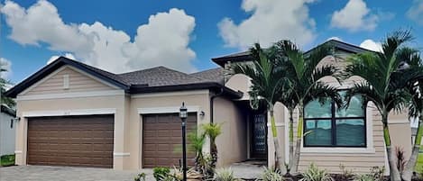 A residential house with two garage doors, a paved driveway, and a well-maintained lawn with palm trees.