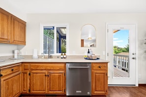 A kitchen with wooden cabinets, a stainless steel dishwasher, a window with a view of the outdoors, and a door leading to a deck. - HillCreek Villa - Spa • Sauna • Cold Plunge • Creekside Shower • Arcade Games (Redding)
