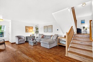 A spacious living room with a wooden floor, a grey sectional sofa, a small armchair, a vintage suitcase, and a staircase with wooden railings. - HillCreek Villa - Spa • Sauna • Cold Plunge • Creekside Shower • Arcade Games (Redding)