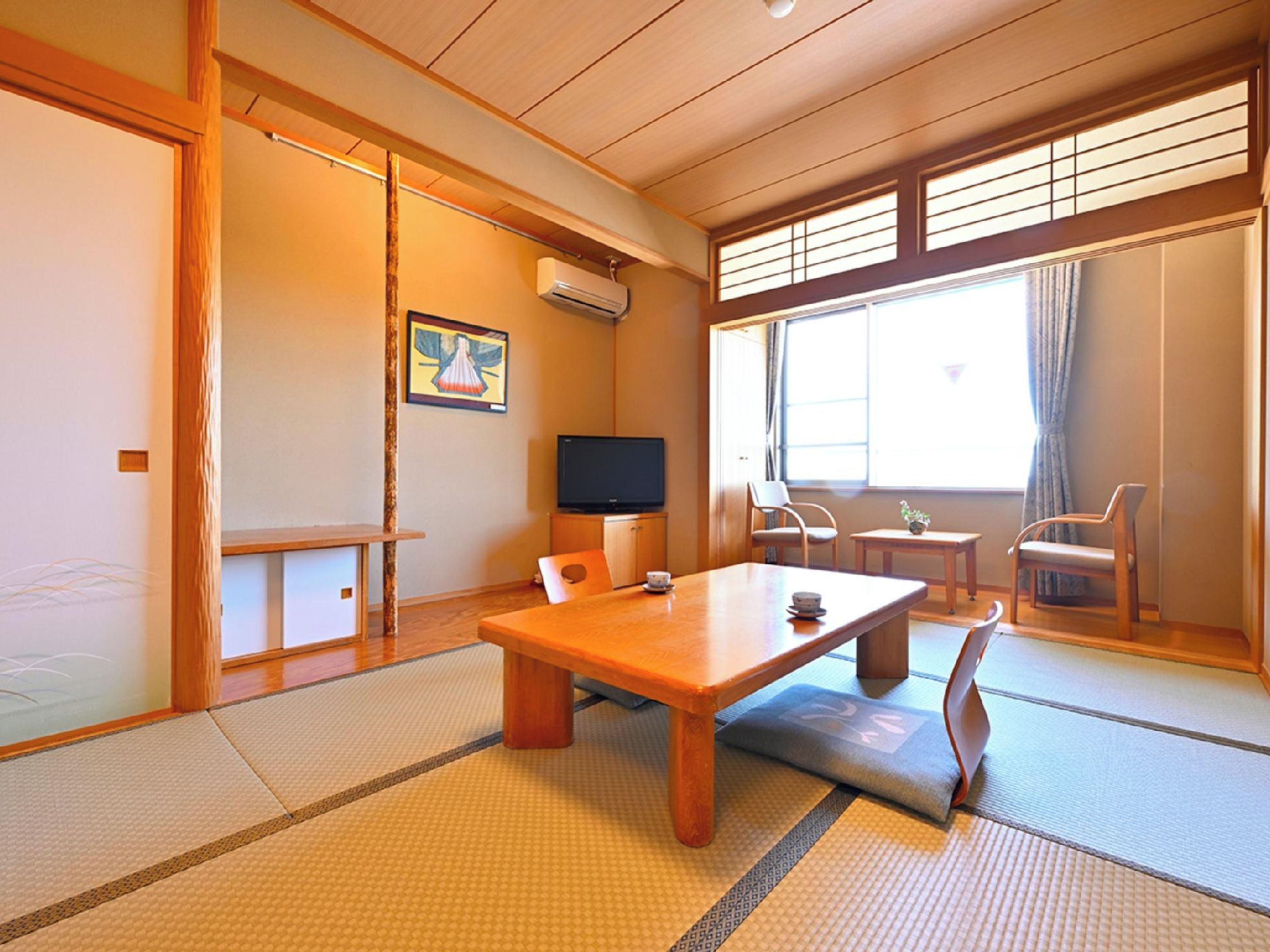 A traditional Japanese room with tatami flooring, a low table, chairs, a television, and a window with curtains.