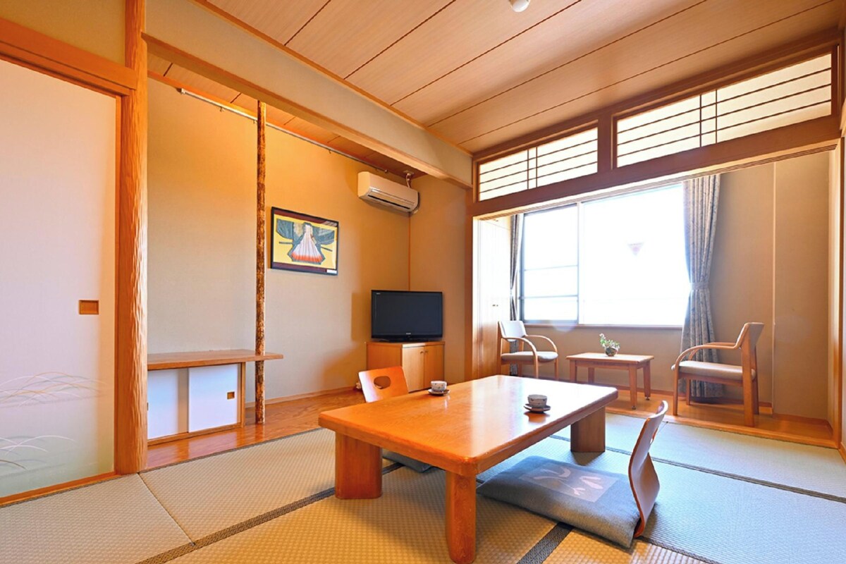Una habitación japonesa tradicional con suelo de tatami, una mesa baja, sillas, un televisor y una ventana con cortinas.