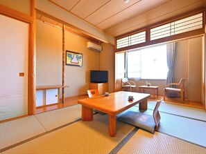Una habitación japonesa tradicional con suelo de tatami, una mesa baja, sillas, un televisor y una ventana con cortinas.