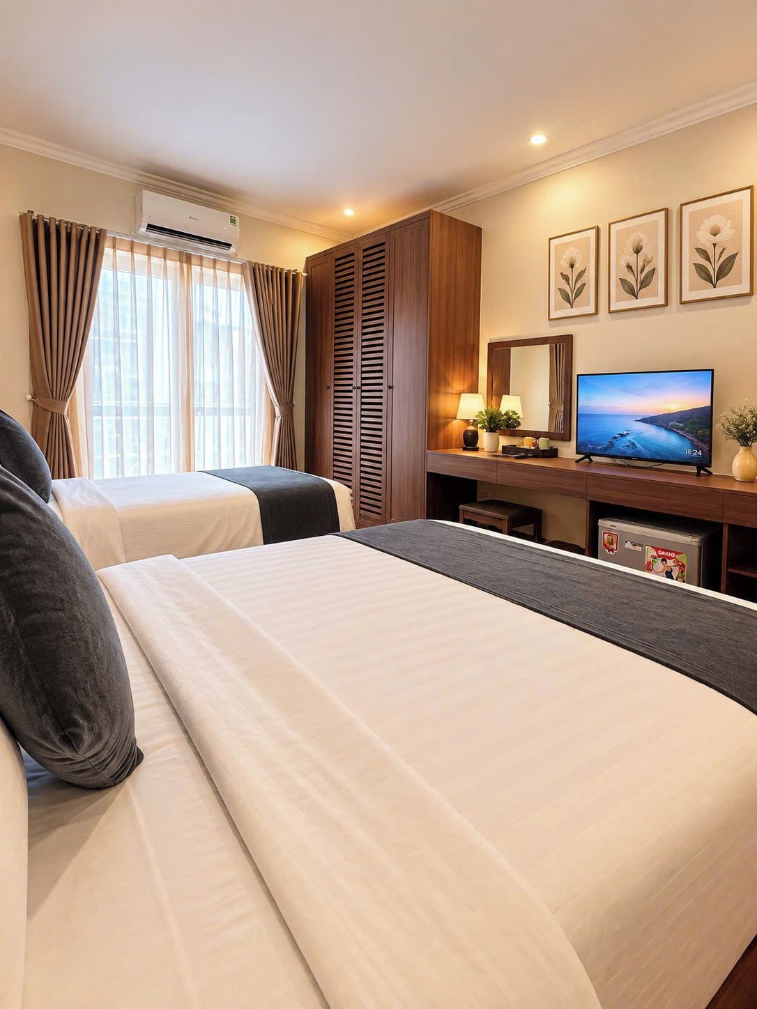 Quarto de hotel com duas camas, televisão, escrivaninha com luminária e janela com cortinas.