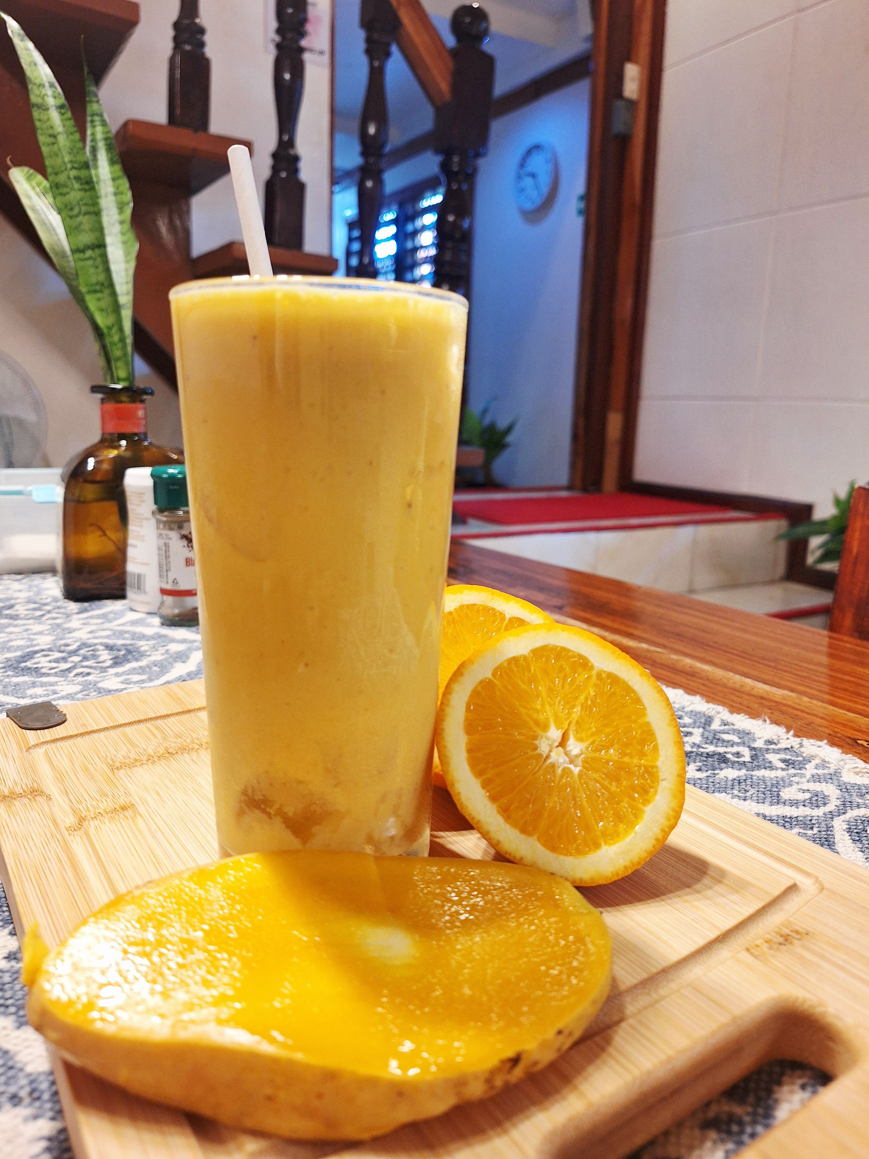 Lasillinen appelsiinimehua puupöydällä, appelsiiniviipale vieressä ja oliiviöljypullo taustalla.