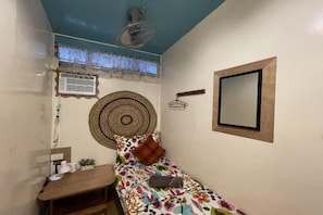 A small, cozy room with a bed, a desk, a fan, an air conditioner, and a window with lace curtains.