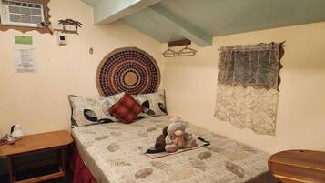 A bedroom with a bed, a bedside table, a wall-mounted heater, a wall clock, and a decorative wall hanging.