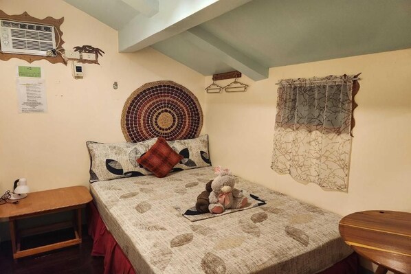 A bedroom with a bed, a bedside table, a wall-mounted heater, a wall clock, and a decorative wall hanging.