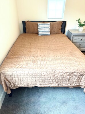 Ein Bett mit gesteppter Decke, ein Nachttisch mit Pflanze und ein Fenster mit Jalousien.
