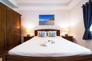 Quarto de hotel com uma cama grande, duas mesas de cabeceira com luminárias, um guarda-roupa de madeira e uma pintura de uma cena de praia na parede.