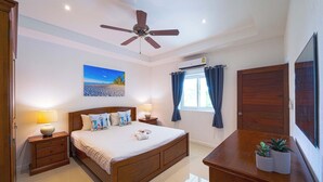 Quarto de hotel com cama, abajures, ventoinha no teto, janela com cortinas, ar-condicionado fixado na parede e televisão de tela plana sobre uma cômoda de madeira.